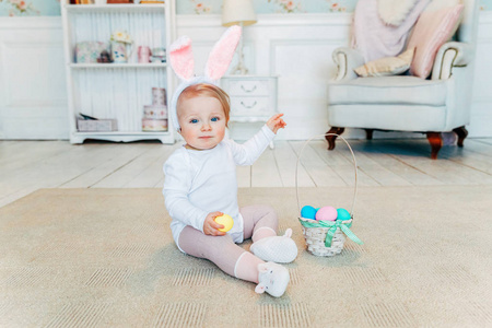 复活节时兔子耳朵的小女孩。 女孩拿着篮子，上面画着鸡蛋，坐在地板上，在家里玩复活节猎蛋的乐趣。 节日庆典春节概念