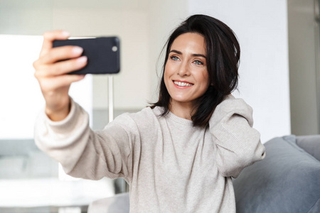 一张30岁的黑发女人坐在明亮的公寓里的沙发上用手机自拍的照片