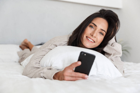 一张30多岁的中年妇女在明亮的房间里躺着白色亚麻布的床上时使用手机的照片