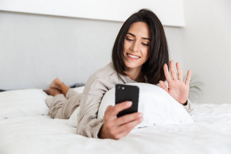 在明亮的房间里躺在床上，穿着白色亚麻布的30岁成年妇女使用手机的照片