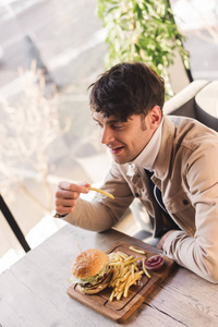 快乐的男人拿着炸薯条，靠近美味的汉堡，在咖啡馆的砧板上
