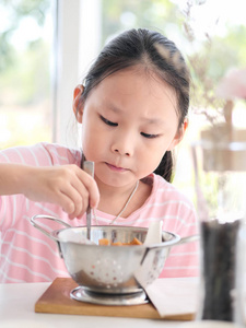 亚洲女孩在家窗户附近的铝碗里吃炸香肠。