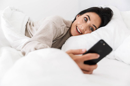一张30岁的的照片，使用智能手机，躺在床上与白色亚麻布在家里。