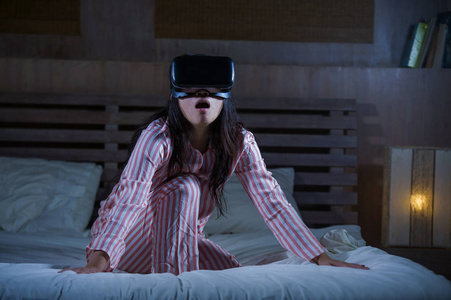年轻的快乐和兴奋的女人玩虚拟现实VR护目镜耳机设备，在床上玩，有3D体验电子游戏，震惊的外观和触摸数字错觉