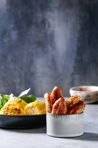 烧烤辛辣的鸡翅放在陶瓷碗里，配上红酱烤甜玉米和浅蓝色桌子上的欧芹包。