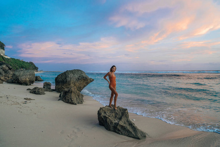 苗条的女孩在泳衣站在大石头在美丽的日落的野生海滩背景