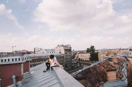 爱的一对夫妇穿着白色连衣裙，戴着帽子的男人在屋顶上散步，拥抱和笑着从城市屋顶上看到积极而真诚的爱情情绪