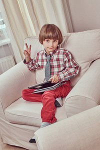 白种人小学生穿着格子衬衫, 领带上有和平的标志, 坐在家里的沙发上拿着一块平板电脑