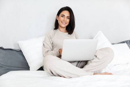在舒适的公寓里，30岁的快乐女人坐在床上，穿着白色亚麻布，用笔记本电脑拍照