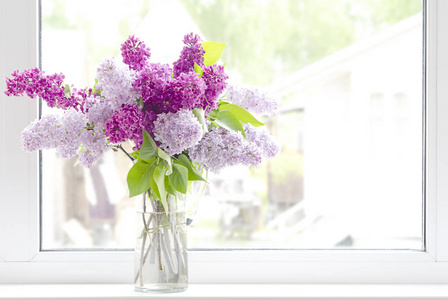 夏天，在阳光明媚的天气里，一小束紫色紫丁香枝在玻璃装饰器中的静物被窗户安装在乡下的一个白色窗台上。