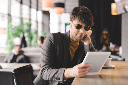 快乐的年轻人戴着太阳镜，坐在酒吧柜台旁，拿着数字平板电脑，端着咖啡