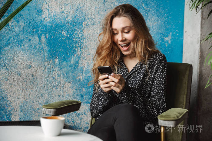 图为美丽的年轻美女坐在室内咖啡厅用手机聊天