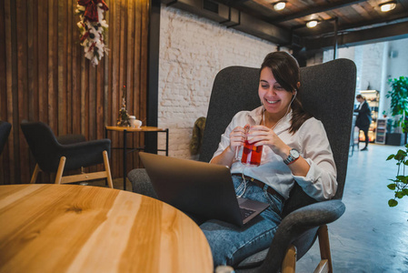 女人坐在有笔记本电脑的咖啡馆里的大椅子上。 用耳机听音乐。 喝水果茶。 自由职业概念