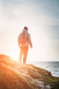 带着背包的男人旅行者站在草丛中的岩石悬崖上, 在日落时看着海景, 自由冒险