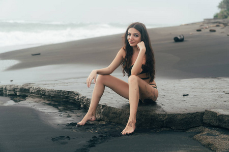 美丽的女孩与长发泳装坐姿和摆姿势在黑色沙滩