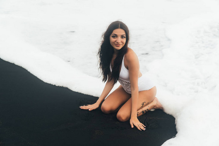 美丽的微笑的女孩在白色泳装选址在黑色的沙滩在海浪的海洋