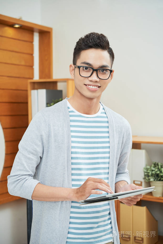 戴眼镜的休闲青年男子微笑着站在办公室里拿着触摸板照片-正版商用