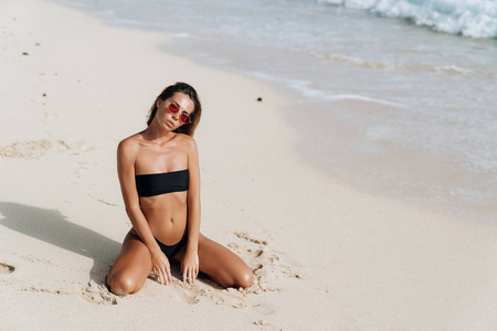 晒黑的女孩在黑色泳衣和太阳镜摆姿势在沙滩上