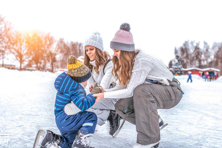年轻的母亲两个女人女友抚养儿子男孩 23年, 在冬季城市公园溜冰场溜冰。概念第一课滑冰支持, 帮助家长学习运动和积极的生活
