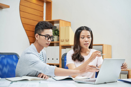 现代亚洲男人和女人看着笔记本电脑，在光办公室的桌子上讨论