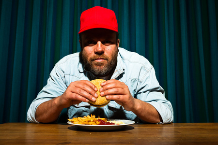 吃快餐餐大胡子的男人。享受法式炸薯条和汉堡。卡车司机与红章