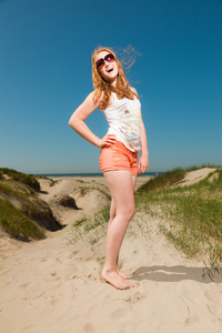 快乐的漂亮女孩与红色的长头发和热的夏天一天享受海滩附近沙丘的太阳镜。清澈的天空