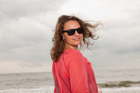 快乐的年轻女人享受海滩附近的户外自然。棕色的头发。身穿粉红色的衬衫和黑色太阳镜。多云的天空