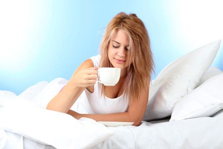 年轻漂亮的女人在床上与杯咖啡在蓝色背景