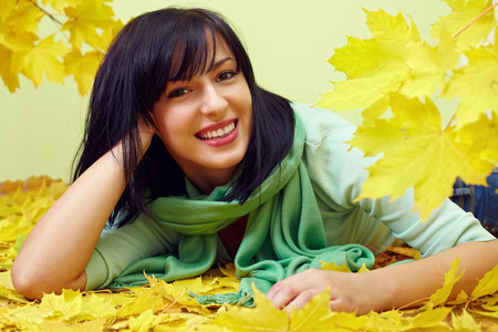 有吸引力微笑着深色头发的女人躺在黄色的落叶，戴着绿色的围巾
