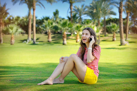 美丽的年轻女人坐在绿色的草坪上，用手机说话