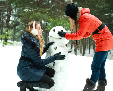 快乐两个漂亮的女孩冬季一天的建设在松林中的雪人