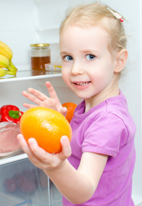 打开冰箱背景上持有橙色快乐微笑的小女孩