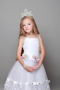 戴着一顶王冠和白的可爱小女孩打扮可爱的微笑和冒充为灰色的背景上的摄像头