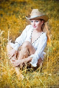 坐在干草 stack.very 可爱金发女人坐在室外黄色在草地上，戴着帽子上的帽子和白色衬衫的时尚肖像女人