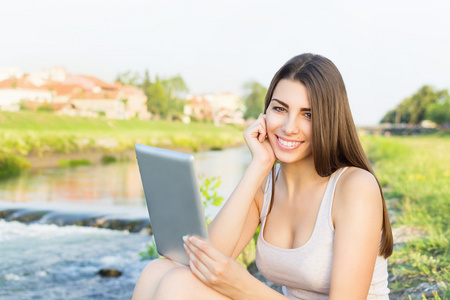 美丽的年轻女子坐在河边使用平板电脑
