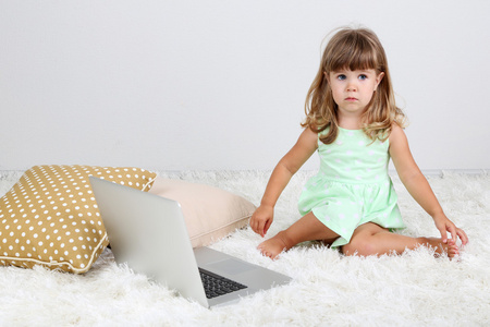 可爱小姑娘坐在地毯上用灰色的背景上的笔记本电脑