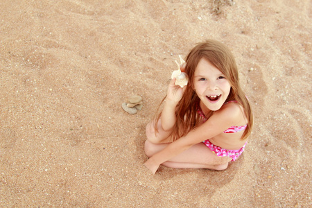 欧洲微笑的小女孩，在坐在海砂在炎热的夏天一天的背景上粉红色泳装