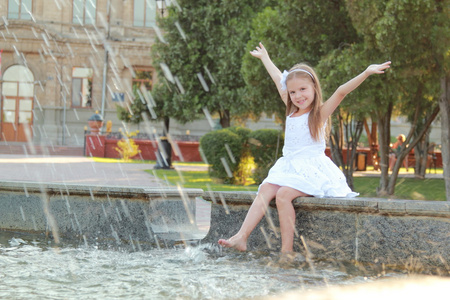 在白色礼服坐在喷水池附近和在夏天主题微笑开朗可爱的小女孩