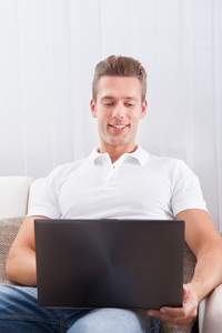 快乐的年轻男子坐在沙发上使用的便携式计算机上