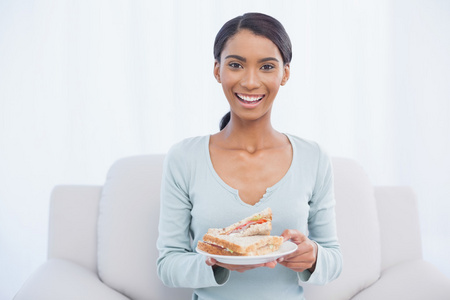 微笑有吸引力的女人坐在舒适的沙发，握着三明治