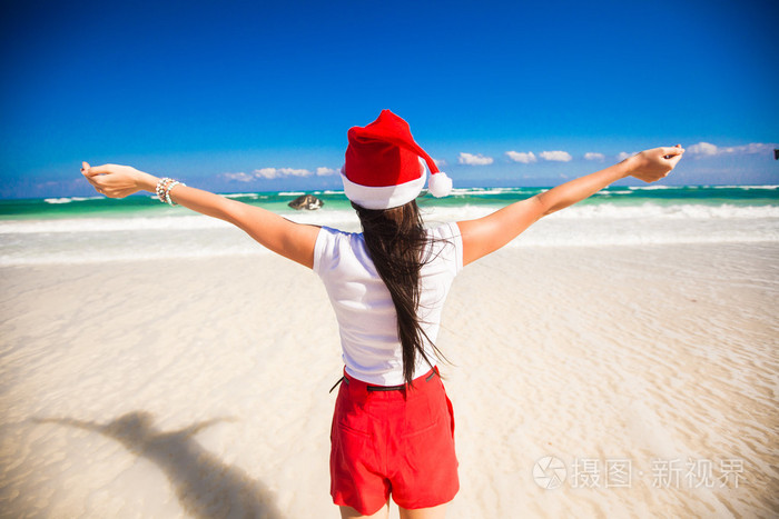 后视图的美丽的女孩在白色沙滩上散步像一只鸟的圣诞老人帽子