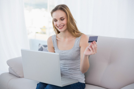 年轻的女人坐在沙发上使用便携式计算机在线购物