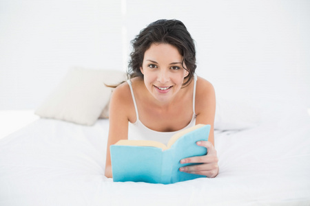 微笑休闲布鲁内特穿着白色睡衣躺在床上读一本书上，
