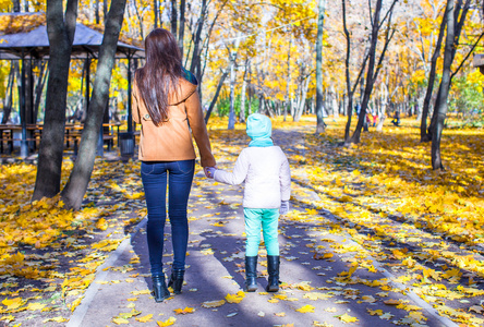 后视图的年轻母亲和她可爱的女儿走在黄色秋天的树林在温暖的阳光灿烂的日子