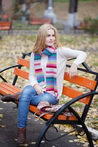 漂亮的金发女孩在秋天的公园里的长椅上坐着的条纹围巾