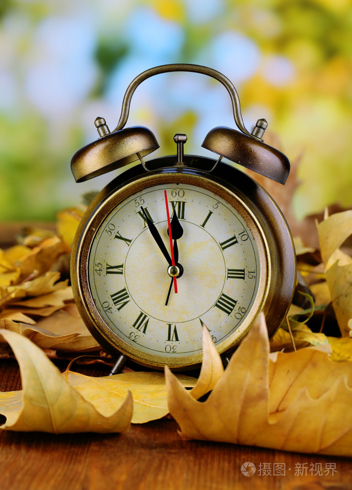 老钟在秋天的落叶,在自然背景上木制的桌子上