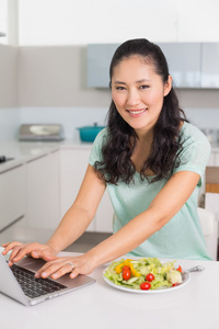 面带笑容的女人，在厨房里吃色拉时使用笔记本电脑