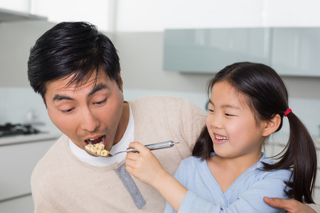 年幼的女儿喂食谷物，父亲在厨房里