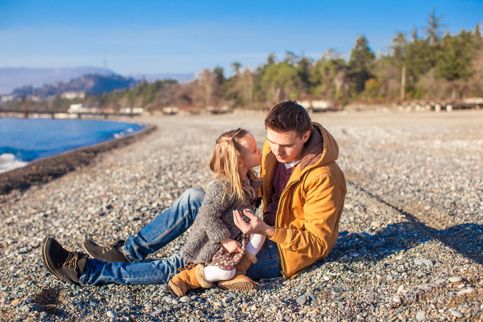 年轻的父亲抱着小女儿亲吻他在海滩上在阳光明媚的日子