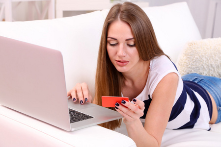 年轻的女人坐在沙发上的笔记本电脑，在她的手，在家里举行信用卡